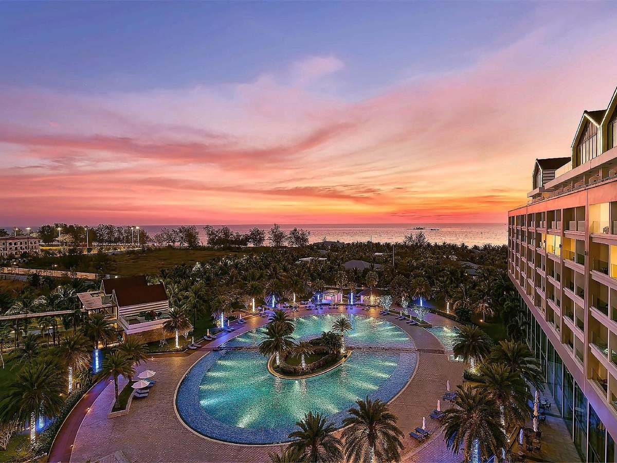 Lagoon pool lit up at dusk around a palm island, beside a tall resort building under a pink sky