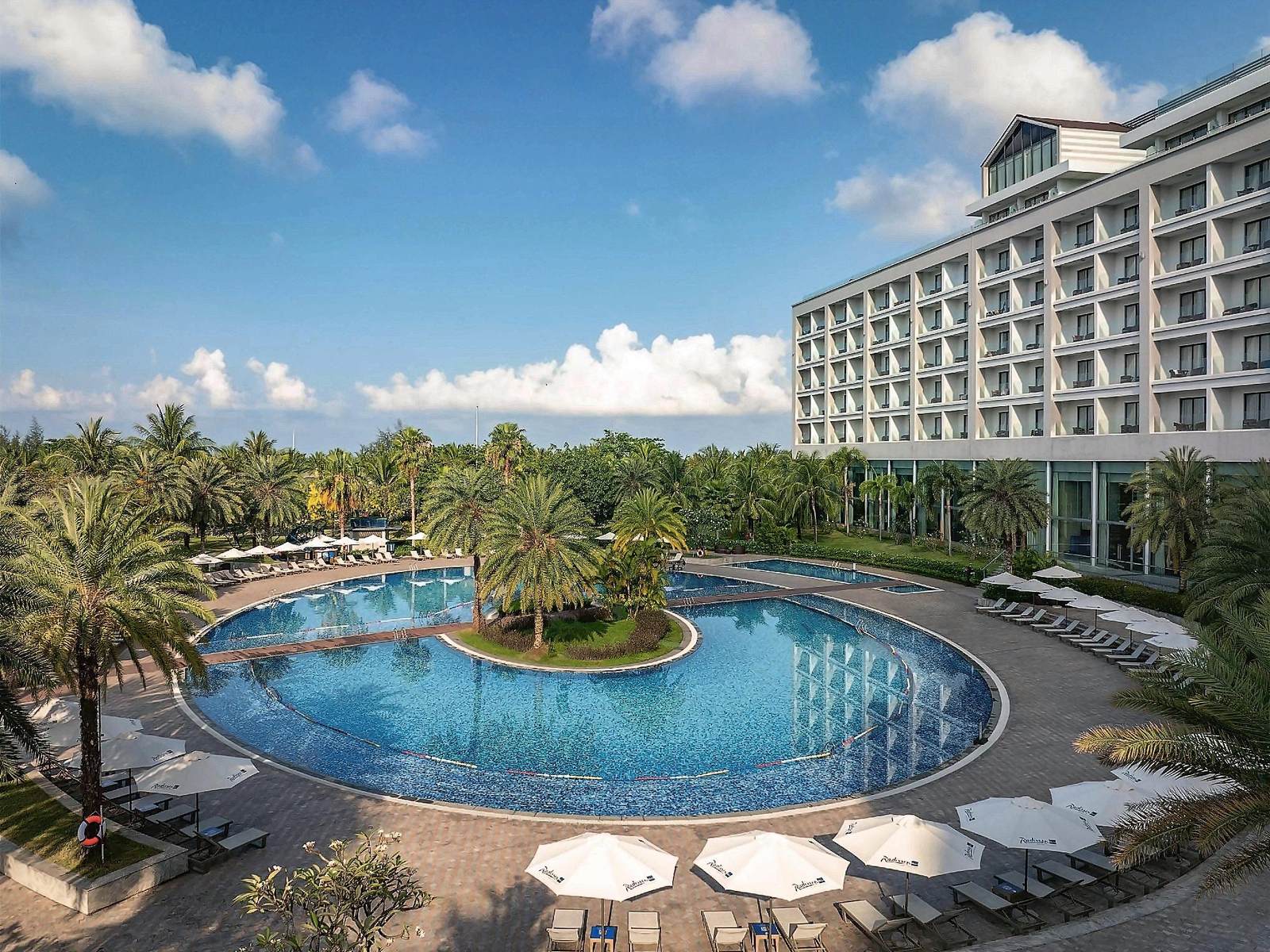 Large curving lagoon pool around a planted island, with loungers, parasols and the white resort building behind