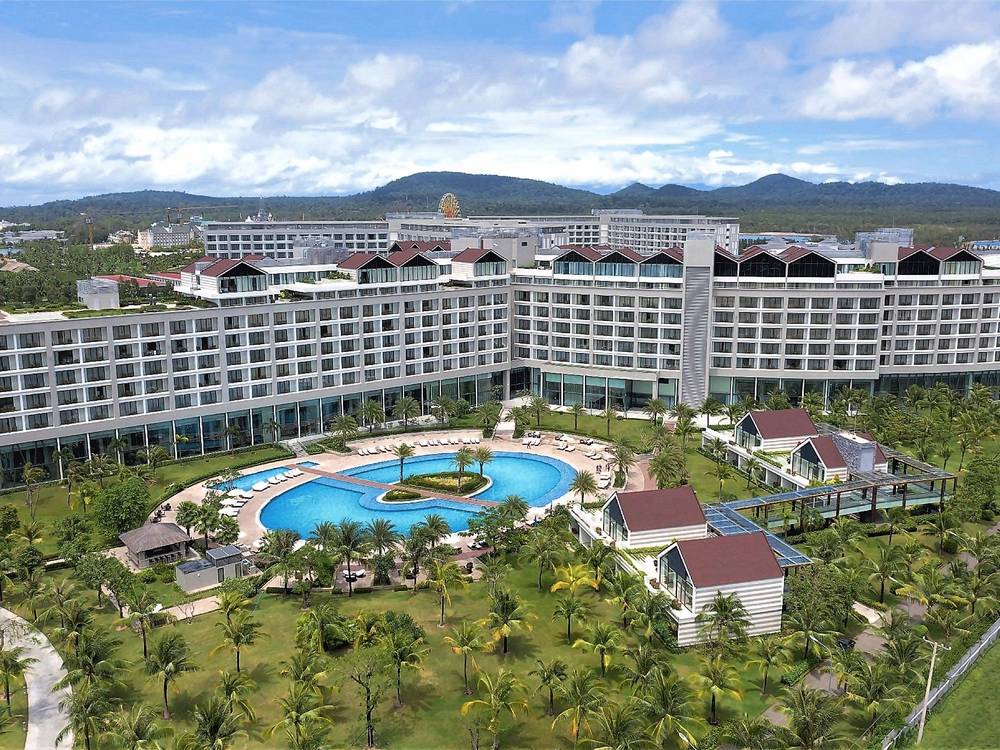 Aerial view of a long white eight-storey resort building with dark roofs, a lagoon pool and palm gardens, with green hills behind