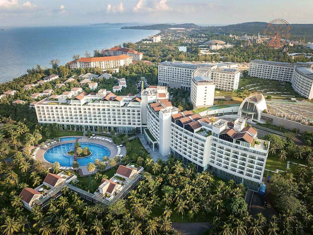 Aerial view of the resort, its lagoon pool and red-roofed buildings among palms, with a Ferris wheel and hills in the distance