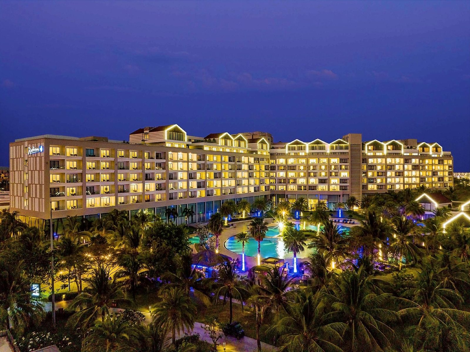 Resort building lit up at dusk above palm gardens and a glowing lagoon pool