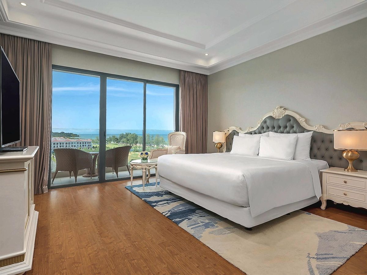Deluxe room with a king bed, carved headboard, wooden floor and a balcony looking over palms towards the sea
