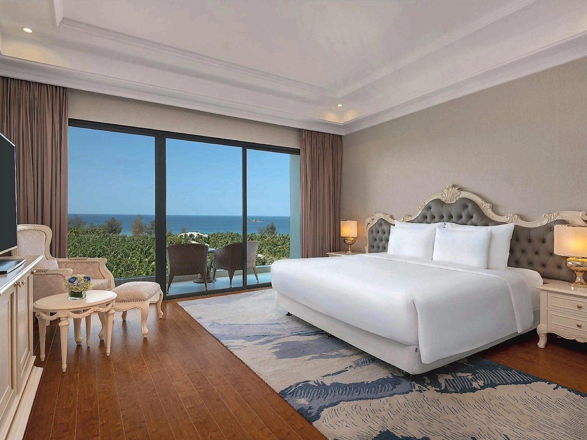 Deluxe room with a king bed, grey tufted headboard, blue rug and glass doors to a balcony facing the sea