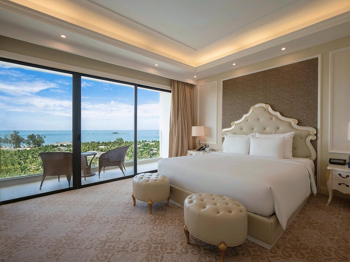 Sea-view suite bedroom with a tufted headboard, upholstered stools and a window over gardens and the sea