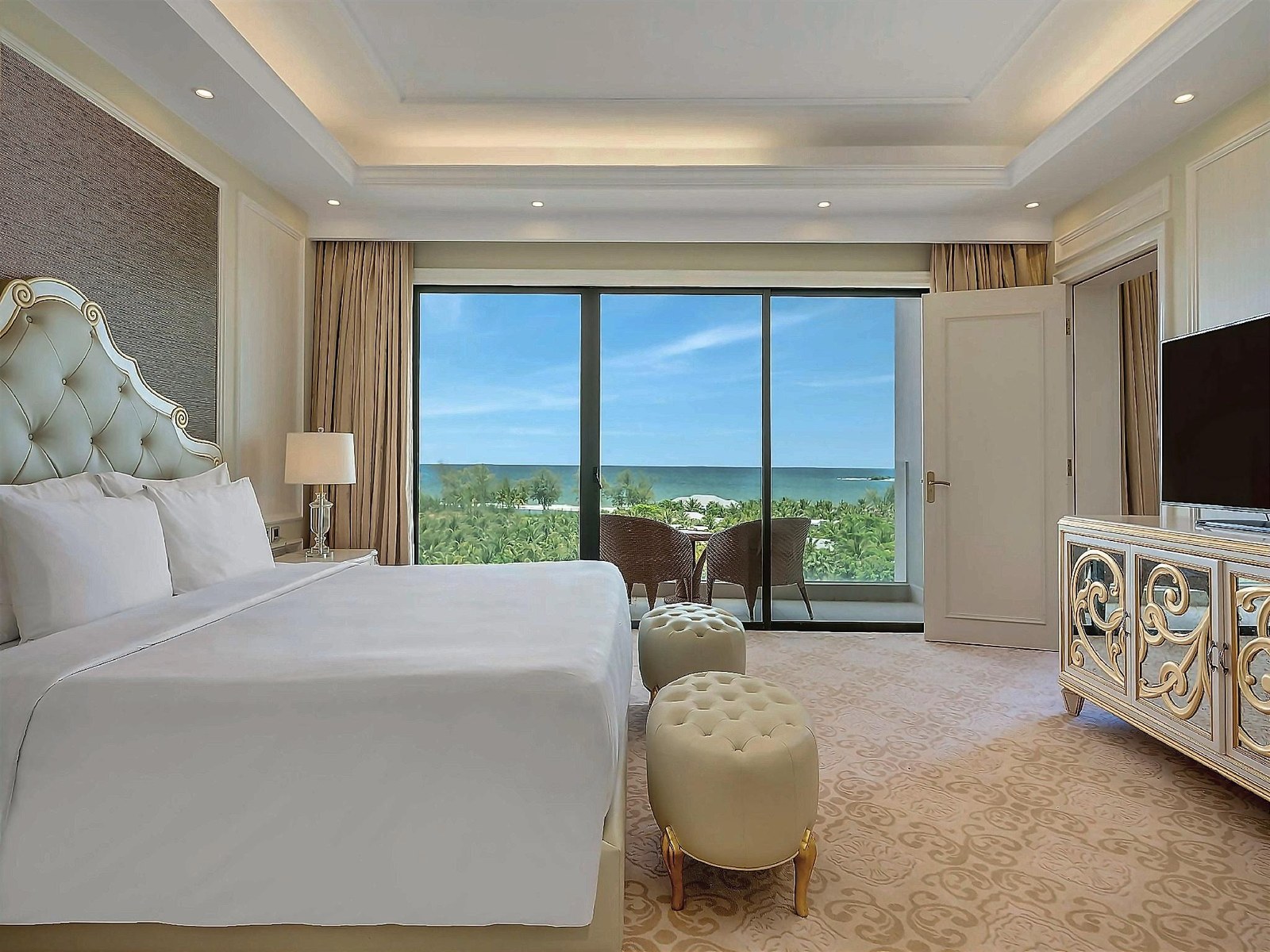 Suite bedroom with a carved white headboard, cream walls and a wide window over palm trees and the sea